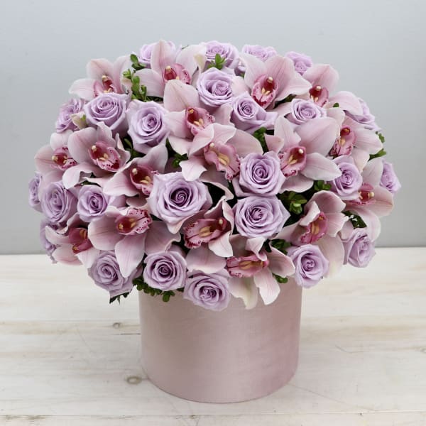 Round arrangement of pale lavender roses and pink orchids in a light pink hat box container.