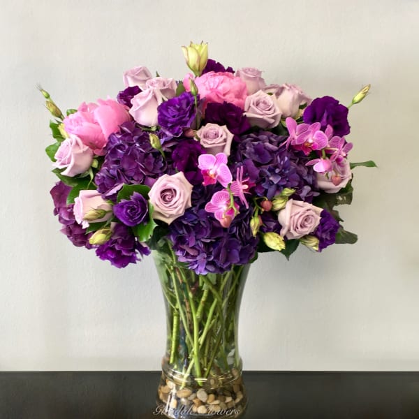 Tall glass vase of purple hydrangeas, lavender roses, and pink blooms in a rounded arrangement