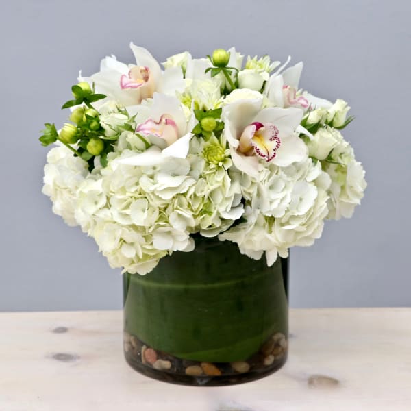 Low round arrangement of white hydrangeas and orchids in a glass cylinder vase with stones.