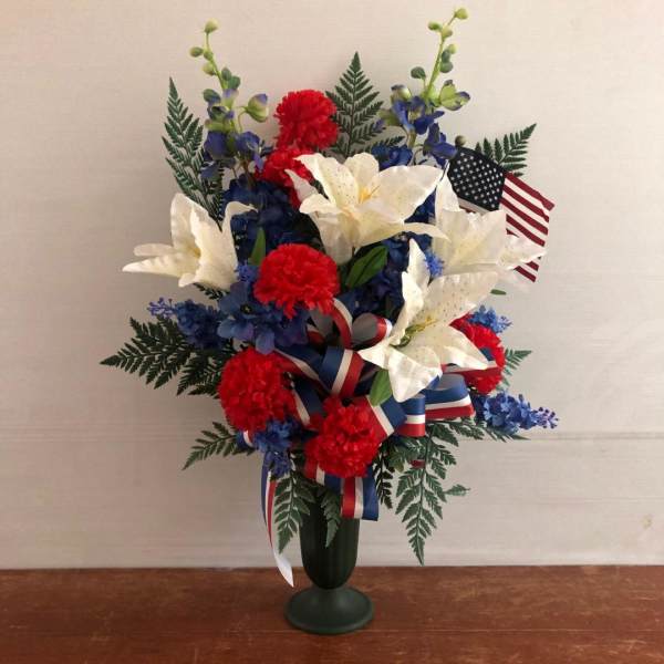 Patriotic arrangement of white lilies and red carnations with blue flowers, ribbon, and a small US flag in a green vase
