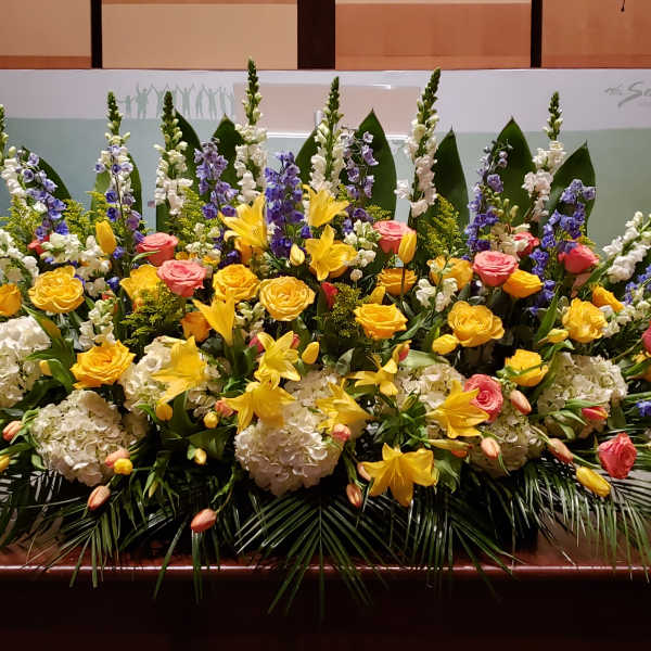 Large stage arrangement of yellow lilies, roses, white hydrangea, and blue delphinium.