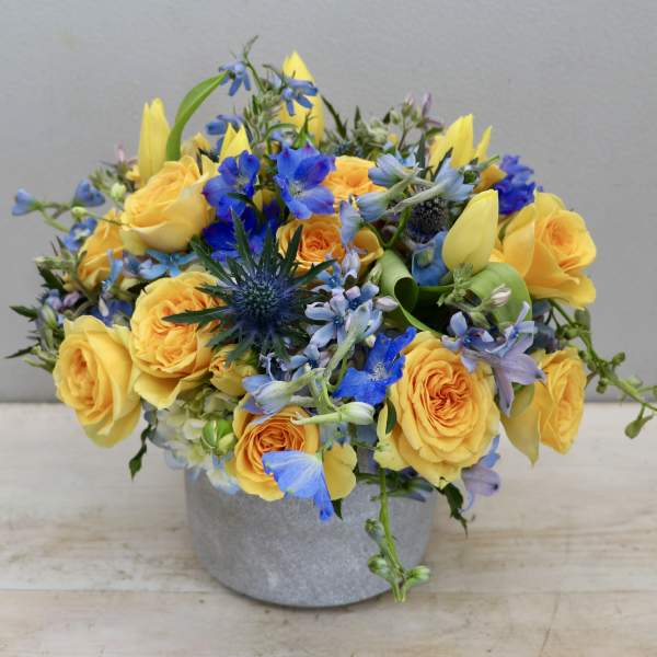 Low arrangement of yellow roses and tulips with blue flowers in a gray pot