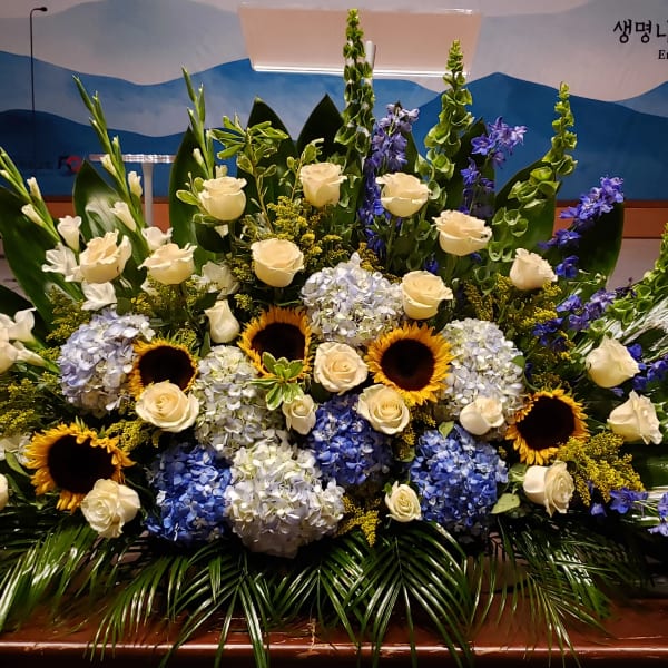Large floral arrangement with white roses, blue hydrangeas, sunflowers, and purple spikes