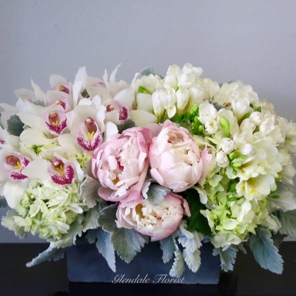 Low rectangular arrangement of white orchids, pale pink peonies, and cream flowers in a dark container