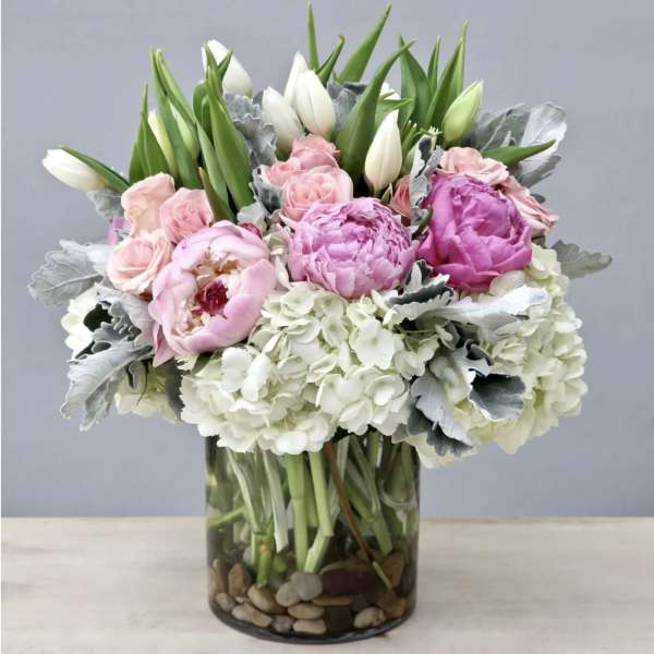 Pink peonies, pale roses, white tulips, and hydrangeas arranged in a clear glass cylinder vase with stones