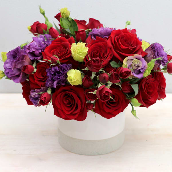 Low arrangement of red roses with purple and light green blooms in a white ceramic vase