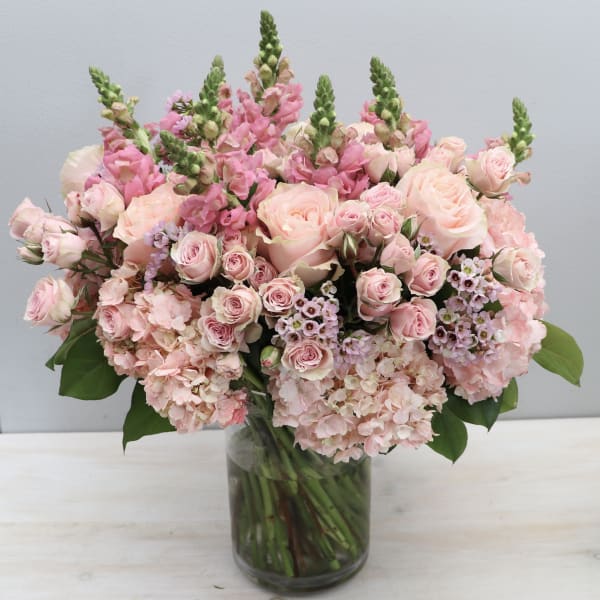 Large arrangement of soft pink roses, snapdragons, and hydrangeas in a clear glass vase.