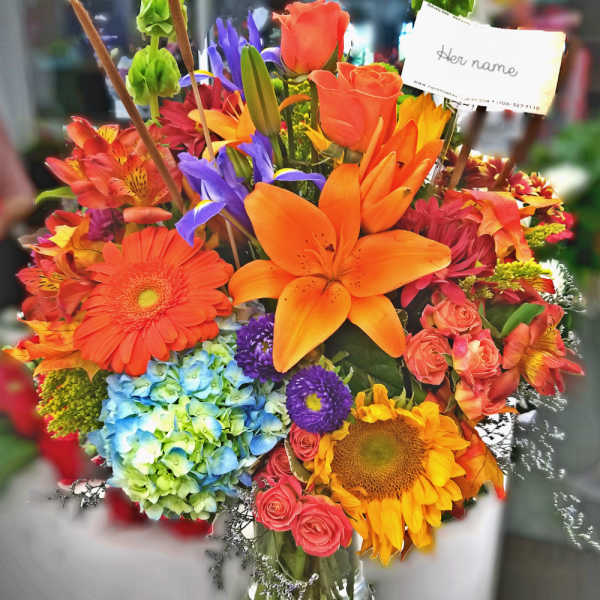 Bright mixed bouquet with orange lilies, roses, sunflowers, and blue hydrangea in a glass vase
