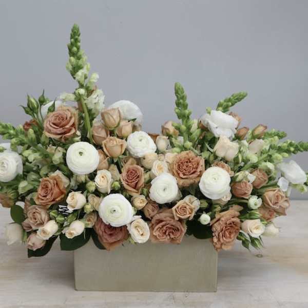 Low rectangular arrangement of white ranunculus and peach roses with snapdragons in a beige container