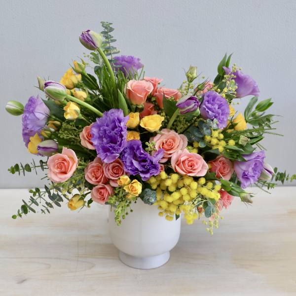 Colorful arrangement of purple lisianthus, peach roses, and yellow blooms in a white ceramic vase