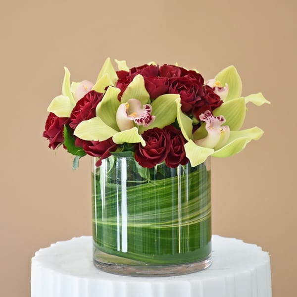 Red roses and pale green orchids arranged in a glass cylinder vase.