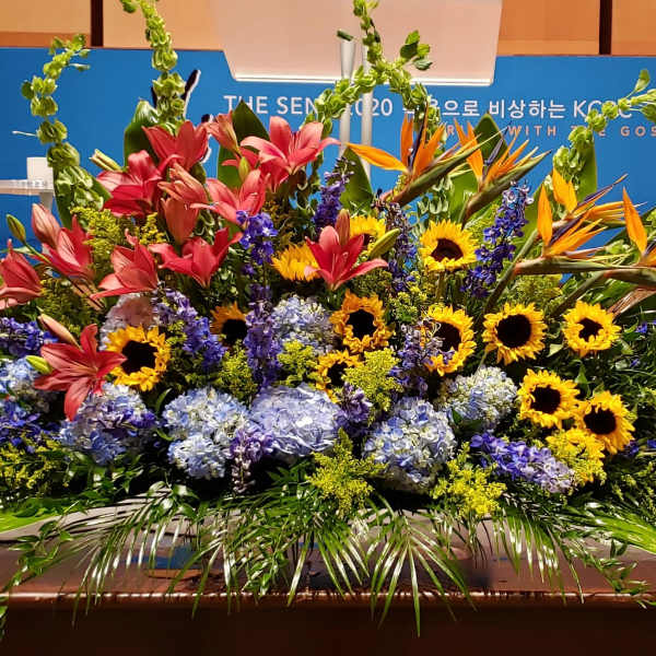 Large stage arrangement with sunflowers, pink lilies, blue hydrangeas, and orange birds of paradise.