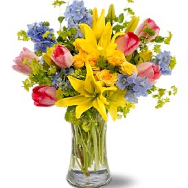 Mixed bouquet of yellow lilies, pink tulips, and blue flowers in a glass vase