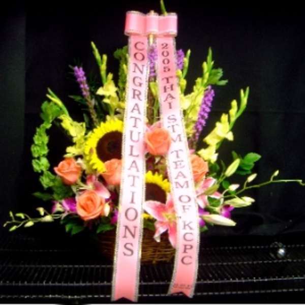 Basket arrangement with sunflowers, pink roses and lilies, accented by tall blooms and pink congratulations ribbons