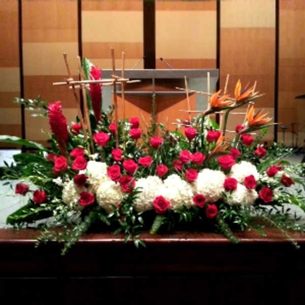 Large altar arrangement of red roses, white mums, and orange tropical flowers with wooden cross accents