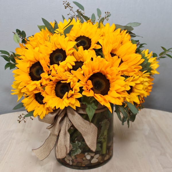 Low round bouquet of yellow sunflowers in a clear glass vase with stones and a burlap bow