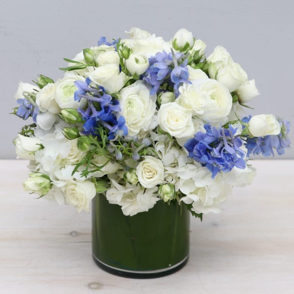 Low round arrangement of white flowers with blue accents in a green cylinder vase