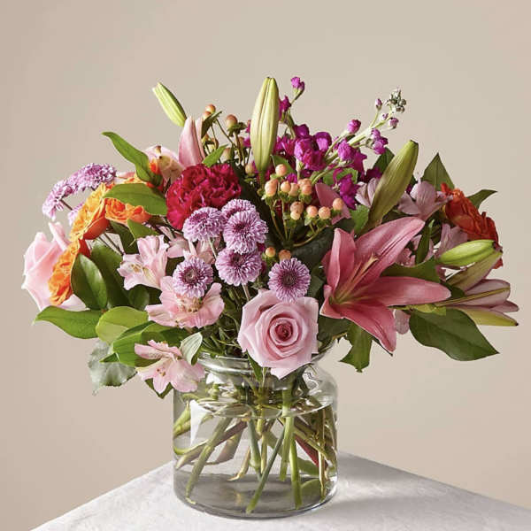 Mixed pink, purple, and orange flower arrangement with roses and lilies in a clear glass vase