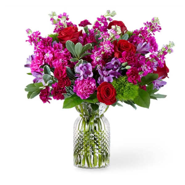 Jewel-tone bouquet of red roses, pink carnations, and purple blooms in a clear glass vase.