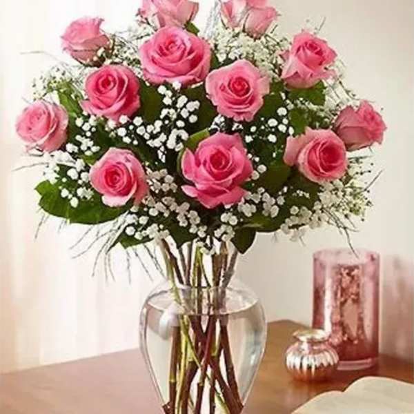 Tall arrangement of pink roses with white filler flowers in a clear glass vase on a wooden table