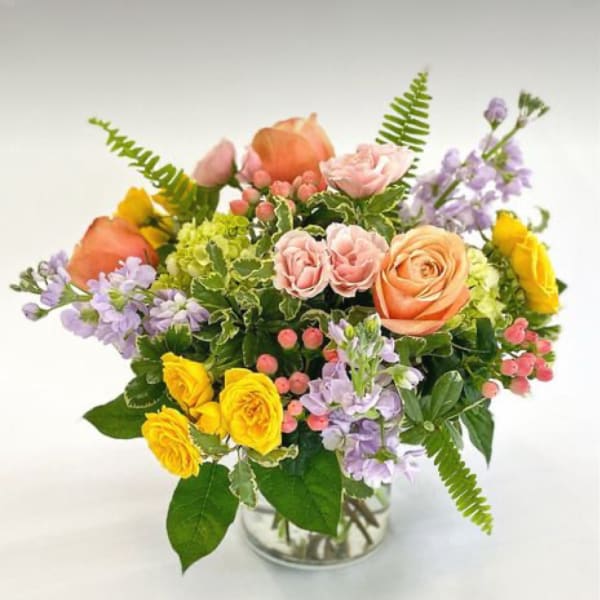 Mixed bouquet of peach and yellow roses, pink spray roses, and lavender blooms in a clear glass vase