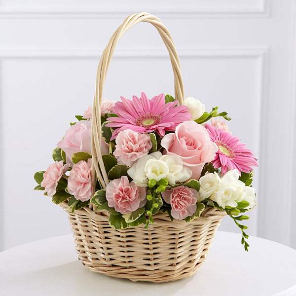 Pastel pink roses, gerbera daisies, and carnations arranged with white blooms in a woven basket.