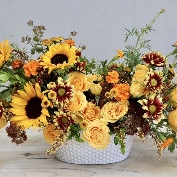 Low oval arrangement of yellow sunflowers, roses, and orange blooms in a white textured ceramic bowl