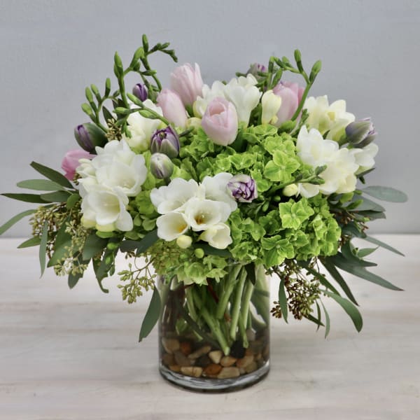 Compact arrangement of pink tulips, white freesias, and green hydrangeas in a clear glass vase with stones.
