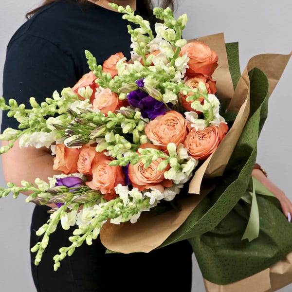 Large hand-tied bouquet of peach roses, white snapdragons, and purple flowers wrapped in kraft paper.