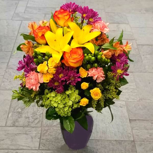 Bright mixed bouquet with yellow lilies, orange roses, and purple daisies in a purple vase
