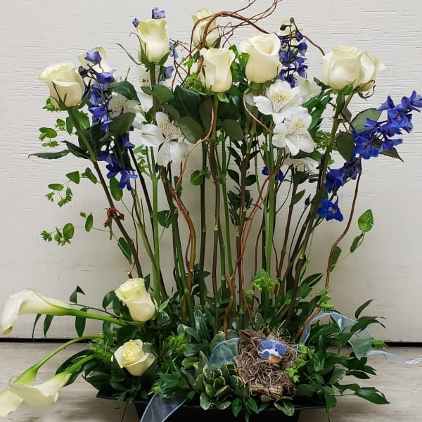 Tall arrangement of white roses, calla lilies, and blue flowers with a small bird nest in a low tray