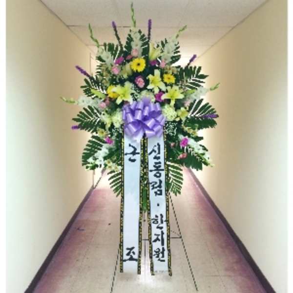 Tall standing spray with yellow lilies and daisies, pink blooms, and a purple bow on an easel with Korean ribbons