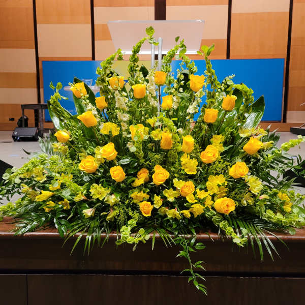 Large fan-shaped arrangement of yellow roses and flowers with lush greenery on a stage front.
