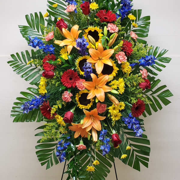 Tall standing spray of orange lilies, sunflowers, red roses, and mixed bright flowers on an easel.