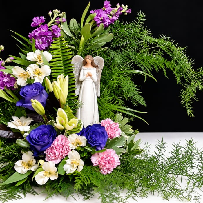 Floral arrangement with an angel figurine, purple roses, pink carnations, and white lilies