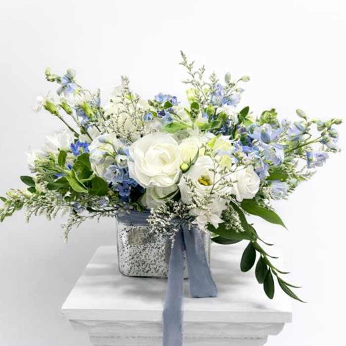 Low white and blue flower arrangement with roses and delphinium in a silver cube vase with blue ribbon