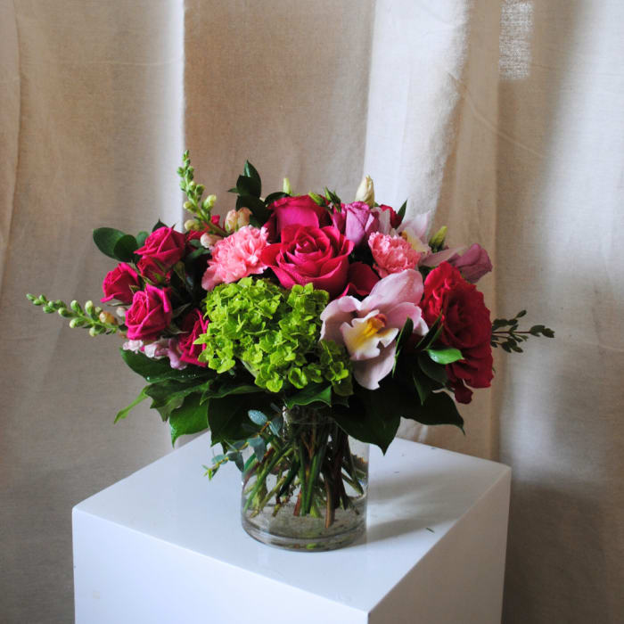Compact arrangement of hot pink roses, pink carnations, and green hydrangea in a clear glass vase