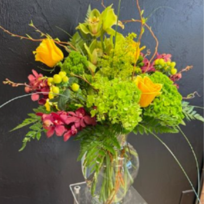 Tall arrangement of orange roses, green hydrangea, and orchids in a clear glass vase