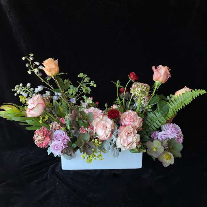 Low pastel arrangement of peach roses, lavender flowers, and green accents in a white rectangular vase