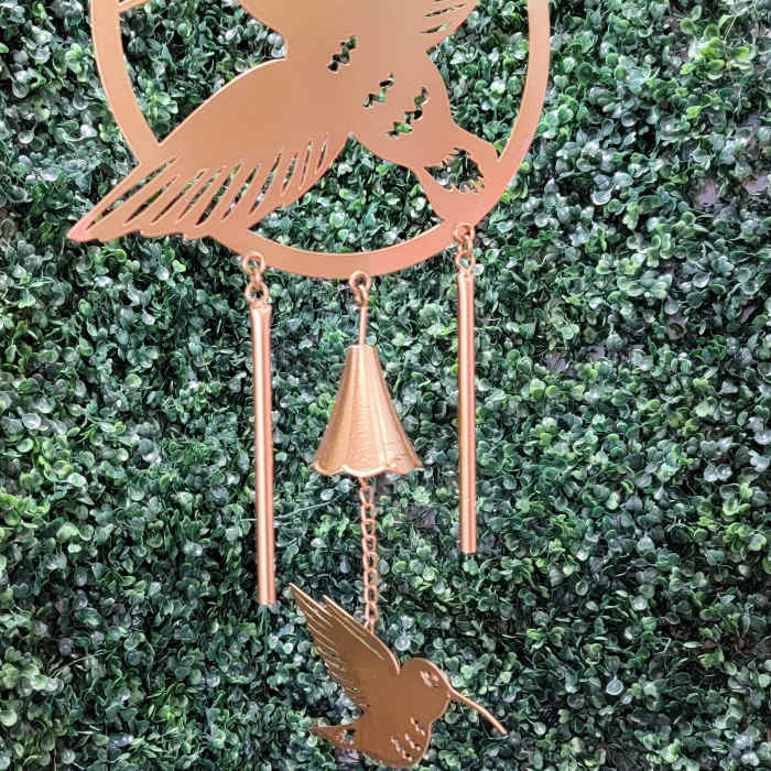 Copper hummingbird wind chime hanging against a green hedge
