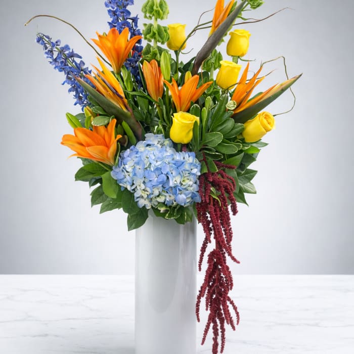 Tall mixed bouquet in a white vase with orange lilies, yellow roses, and blue flowers