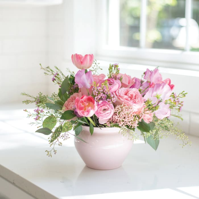 Low pink arrangement of tulips, roses, and ranunculus in a pale pink ceramic bowl