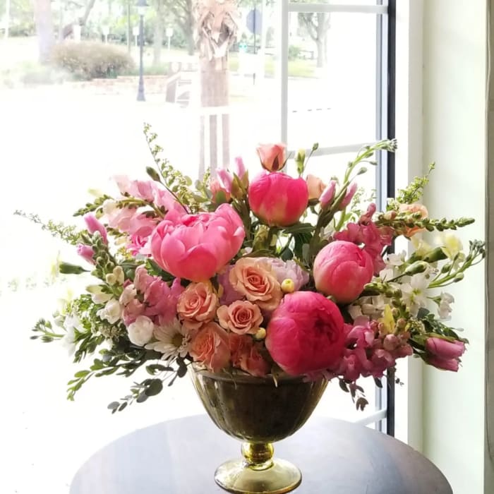 Low gold compote of bright pink peonies, peach roses, and white blooms on a table by a window