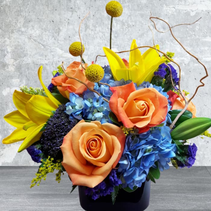 Bright bouquet of orange roses, blue hydrangeas, and yellow lilies in a black vase
