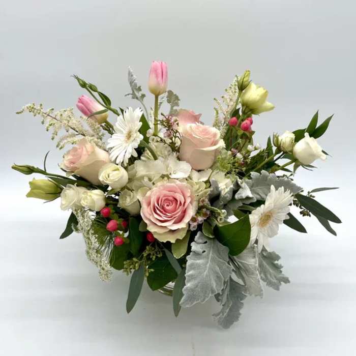 Low arrangement of pink roses, white gerbera daisies, tulips, and silver foliage in a clear glass vase