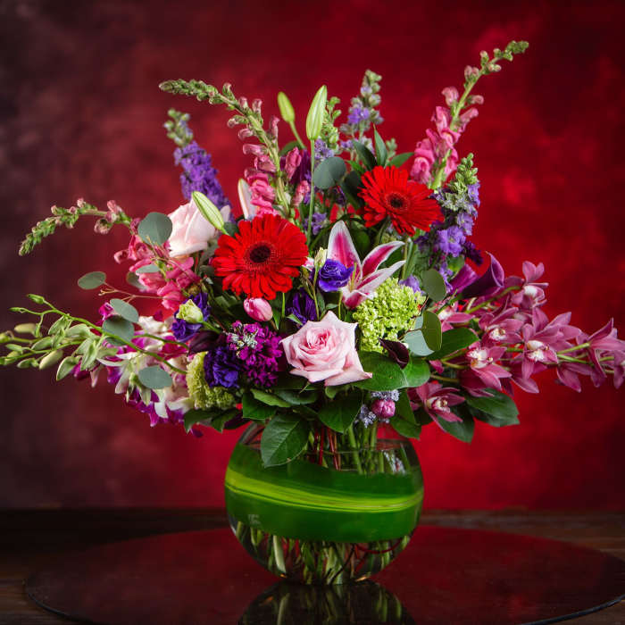 Mixed bouquet with red gerbera daisies, pink roses, and purple blooms in a green glass vase