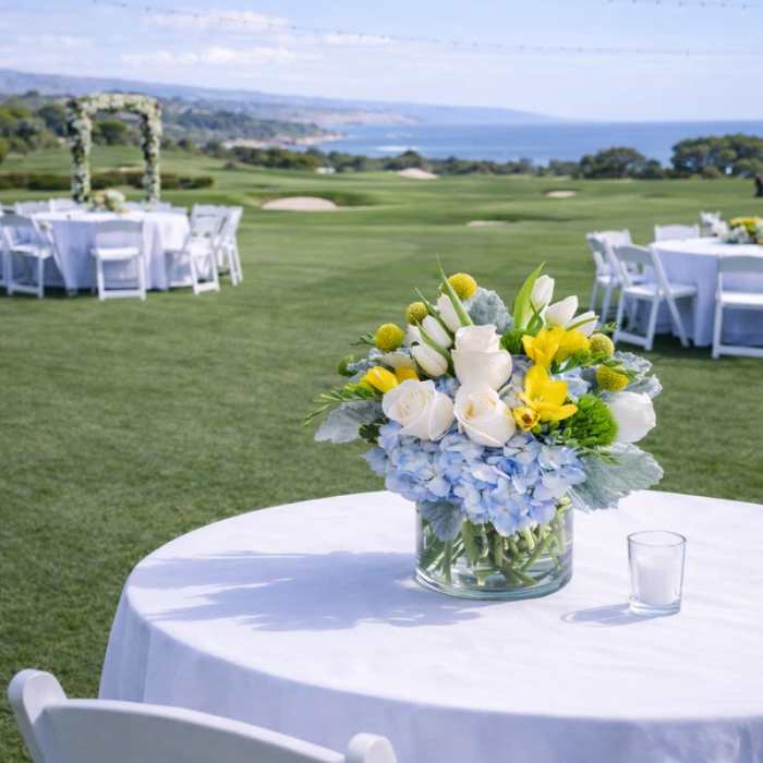 Low blue, white, and yellow flower centerpiece in a clear vase on an outdoor table with white linens