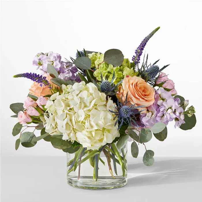 Low glass vase of white hydrangeas, peach roses, and pastel blooms with blue accents.