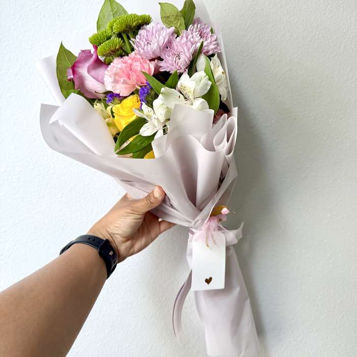 Handheld bouquet of pink, white, and yellow flowers wrapped in pale paper