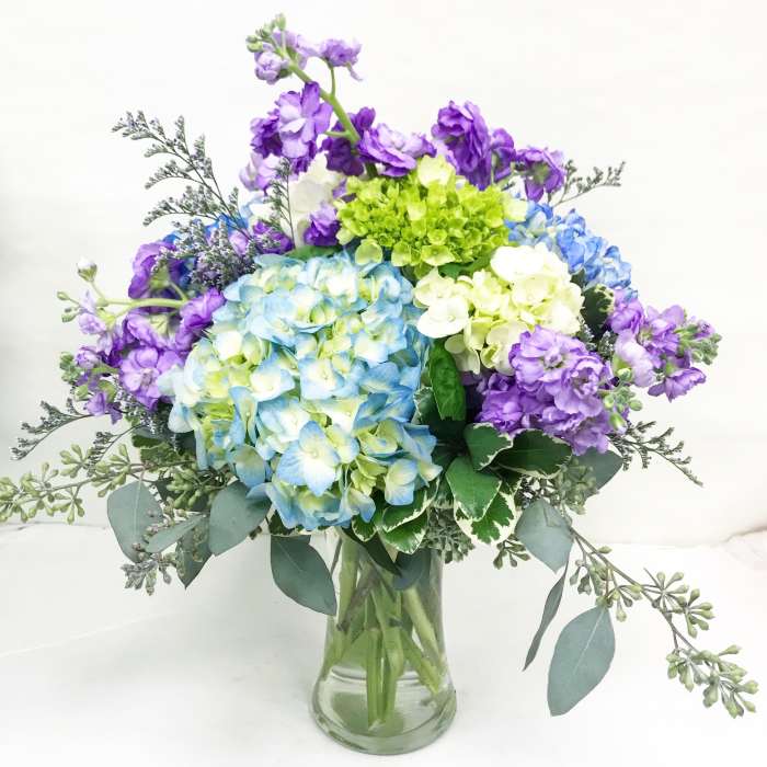 Arrangement of blue and white hydrangeas with purple stock in a clear glass vase